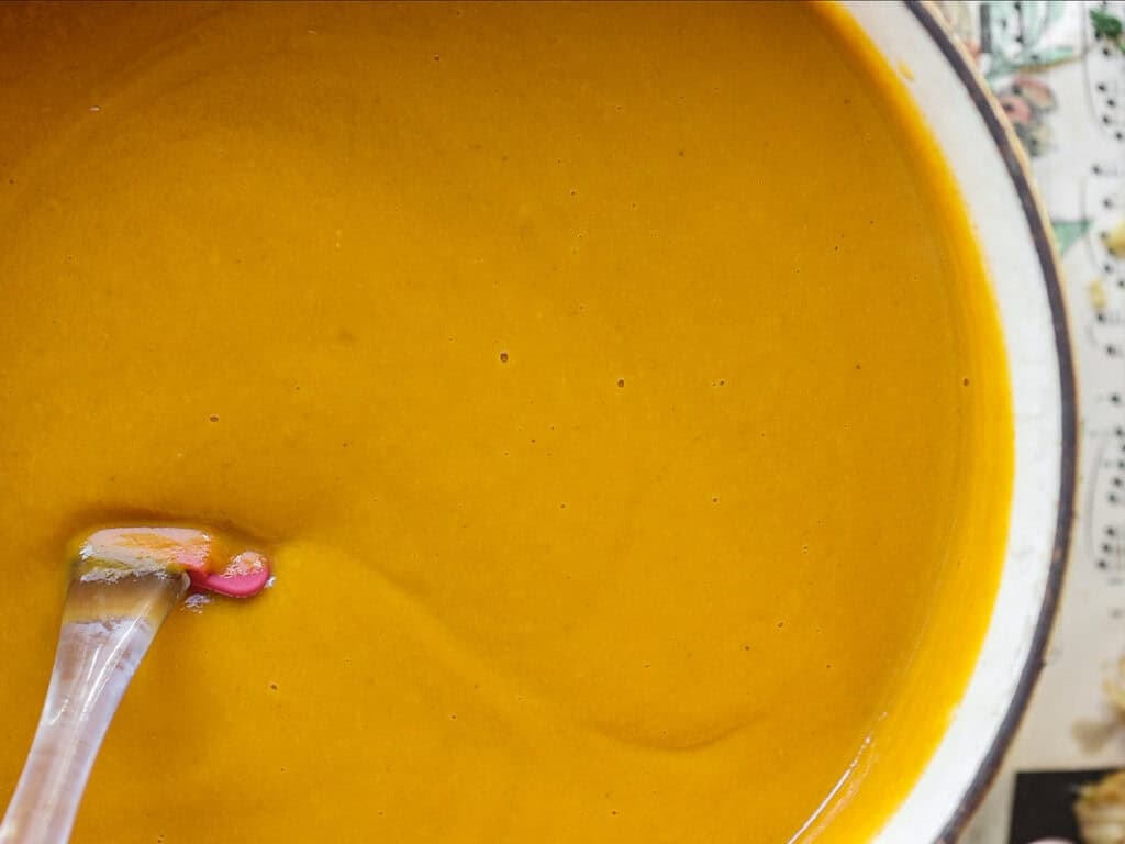 Gochujang and Roasted Carrot Soup - SPICY AND EASY. 15 A close-up of a spoon dipped in a pot of creamy, orange-colored soup. The soup has a smooth texture, and the pot rests on a surface with a faint, patterned design visible on the edges.