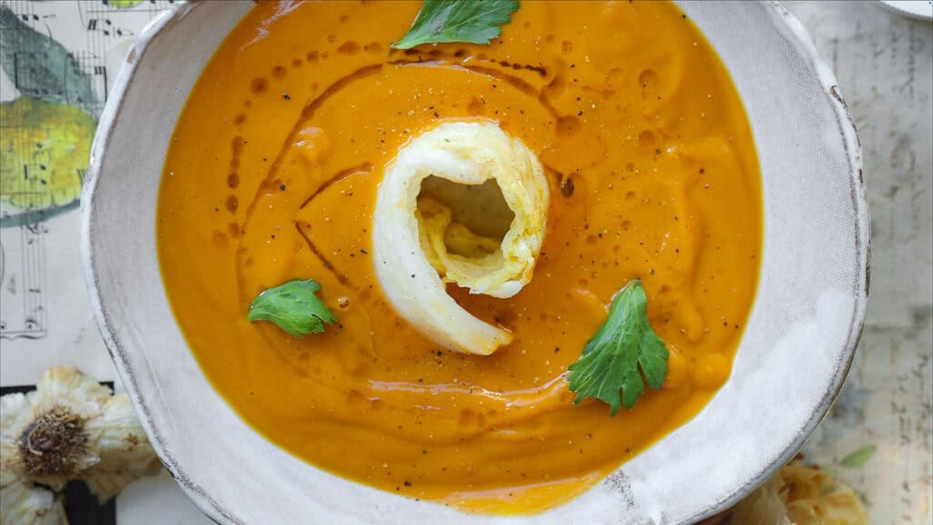 Gochujang and Roasted Carrot Soup - SPICY AND EASY. 1 A bowl of gochujang and roasted carrot soup, garnished with a swirl of cream and fresh parsley leaves. The soup is in a white bowl placed on a patterned surface with hints of garlic visible nearby.