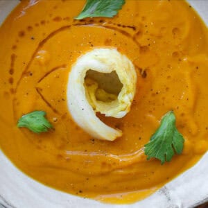 Gochujang and Roasted Carrot Soup - SPICY AND EASY. 19 A bowl of gochujang and roasted carrot soup, garnished with a swirl of cream and fresh celery leaves. The soup is in a white bowl placed on a patterned surface with hints of garlic visible nearby. square