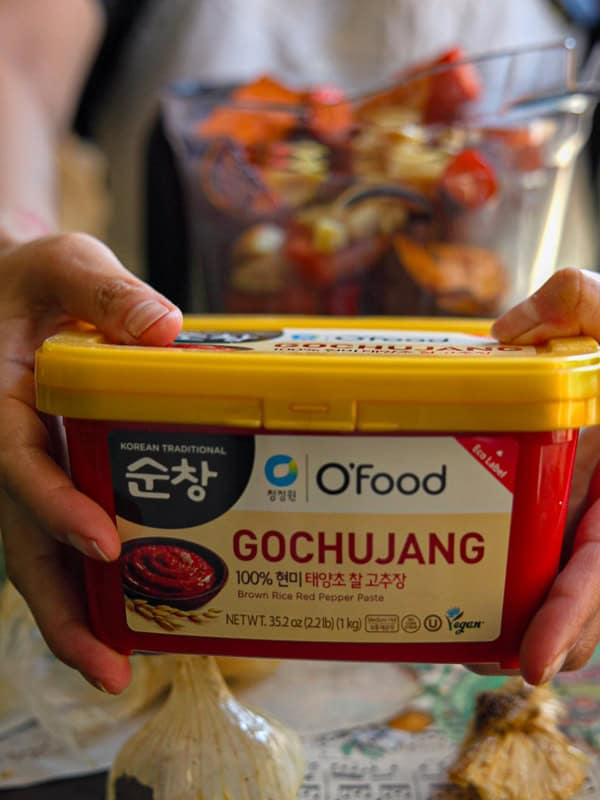 30-Minute Creamy Gochujang Vodka Pasta - Delicious & Dairy-FREE. 6 A person holding a container of OFood Korean Traditional Gochujang, a brown rice red pepper paste, with visible garlic bulbs below. In the background is a glass bowl with chopped vegetables.