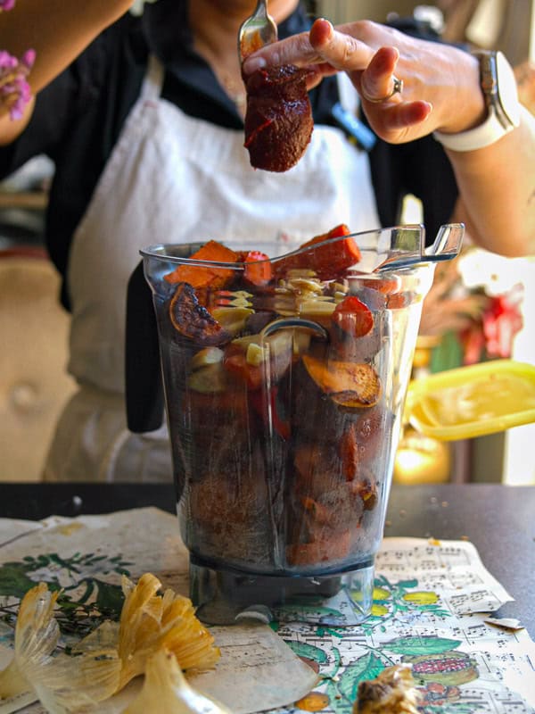 Gochujang and Roasted Carrot Soup - SPICY AND EASY. 12 A person wearing an apron prepares to blend a variety of colorful ingredients including fruits, vegetables, and spices in a blender. They hold a spoon above the blender, suggesting an addition of gochujang. Nearby are corn husks and paper.
