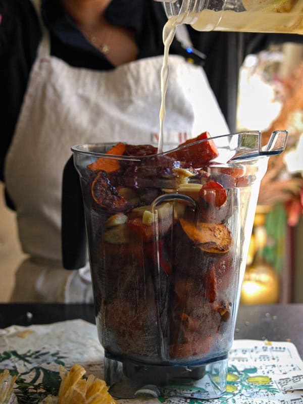 Gochujang and Roasted Carrot Soup - SPICY AND EASY. 13 A person wearing an apron is pouring liquid into a blender filled with various chopped vegetables, including sweet potatoes and bell peppers. The scene suggests meal preparation in a kitchen setting.