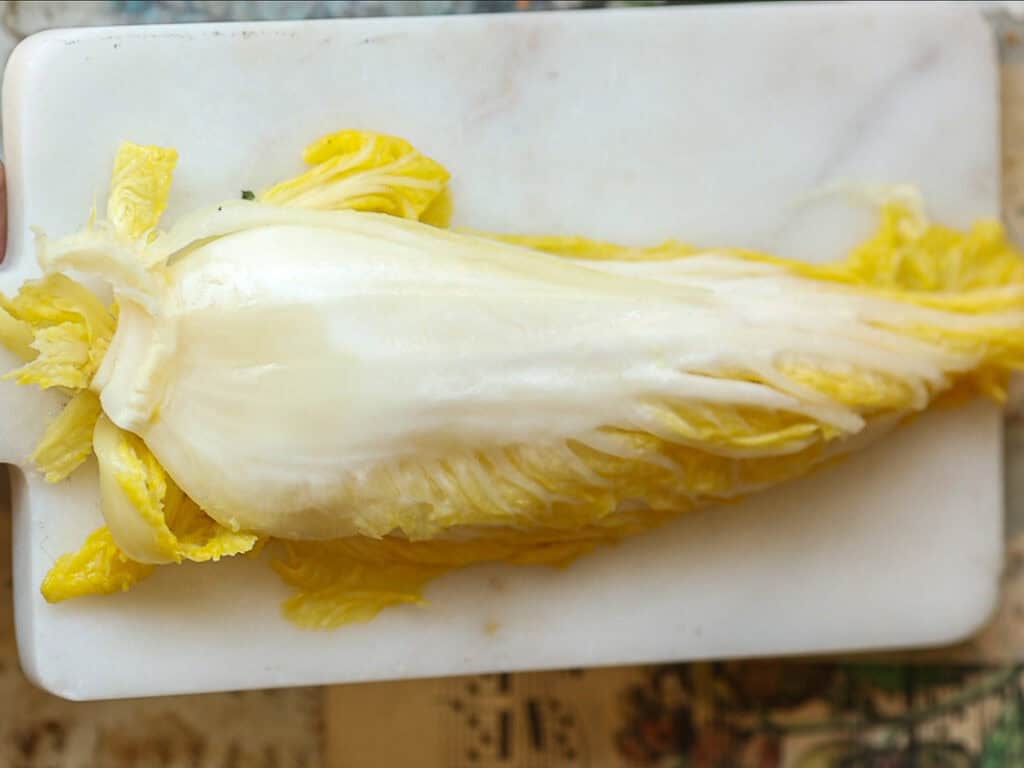 Mom's Easy White Kimchi (Baek-Kimchi) Recipe (백김치). 3 A half of napa cabbage with light yellow and white leaves lies on a white rectangular marble cutting board, showcasing its layers.