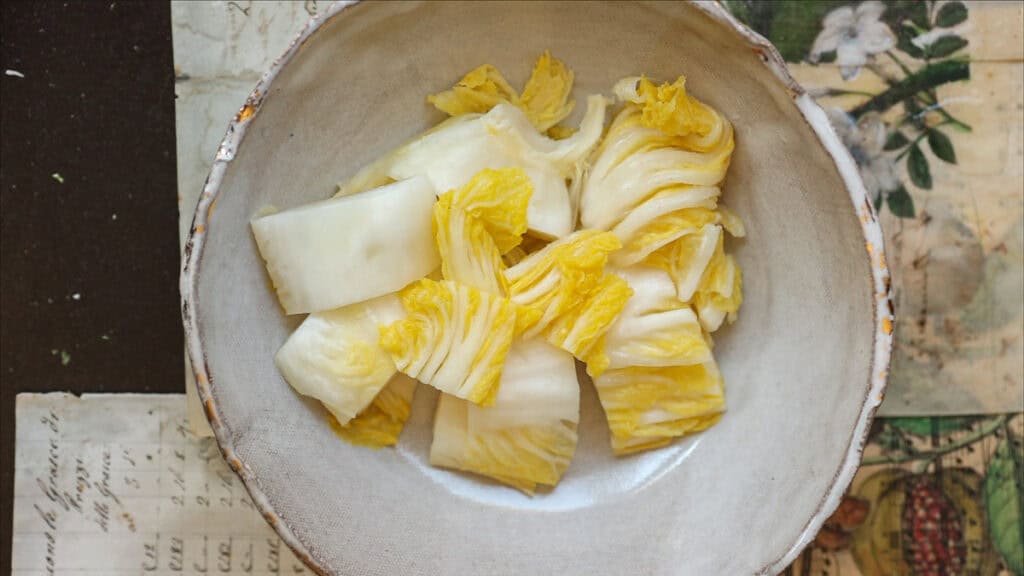 Mom's Easy White Kimchi (Baek-Kimchi) Recipe (백김치). 4 A bowl of Baek-Kimchi, or 백김치, rests on a table adorned with vintage floral and script-patterned papers. The napa cabbage is pale yellow and white, while the bowl exudes a rustic charm, perfectly capturing the simplicity of an easy white kimchi recipe.