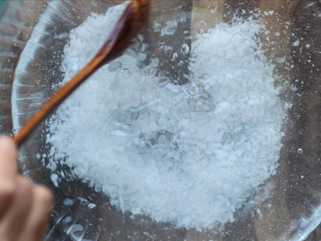 Mom's Easy White Kimchi (Baek-Kimchi) Recipe (백김치). 11 Close-up of a glass bowl with a white substance being mixed by a wooden spoon. The texture is granular and resembles salt surrounded by clear liquid. The bowl is on a wooden surface.