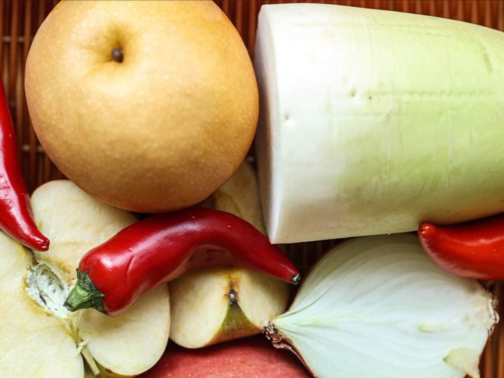 Mom's Easy White Kimchi (Baek-Kimchi) Recipe (백김치). 6 A variety of fresh produce including a whole Asian pear, daikon radish, sliced apples, two red chili peppers, and a cut onion displayed on a wooden surface.