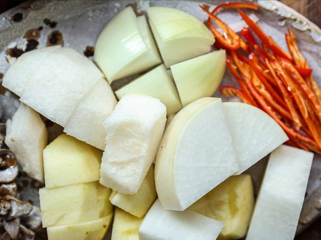 Mom's Easy White Kimchi (Baek-Kimchi) Recipe (백김치). 10 A plate with chopped ingredients including onion quarters, sliced red chili peppers, cubed white radish, and potato chunks. The items are arranged closely together, showcasing a variety of textures and colors.