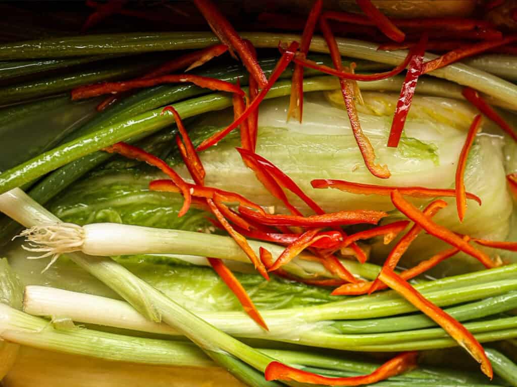 Mom's Easy White Kimchi (Baek-Kimchi) Recipe (백김치). 28 Close-up of fresh vegetables, including green onions, napa cabbage, and thin slices of red chili peppers, arranged in a colorful display.
