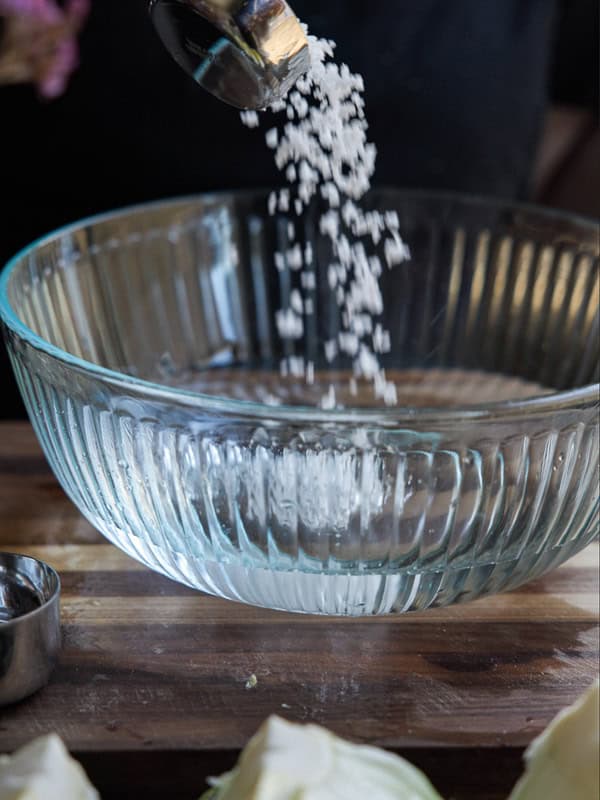 Mom's Easy White Kimchi (Baek-Kimchi) Recipe (백김치). 9 Salt being poured into a clear ribbed glass bowl on a wooden surface, with a metal measuring cup nearby.