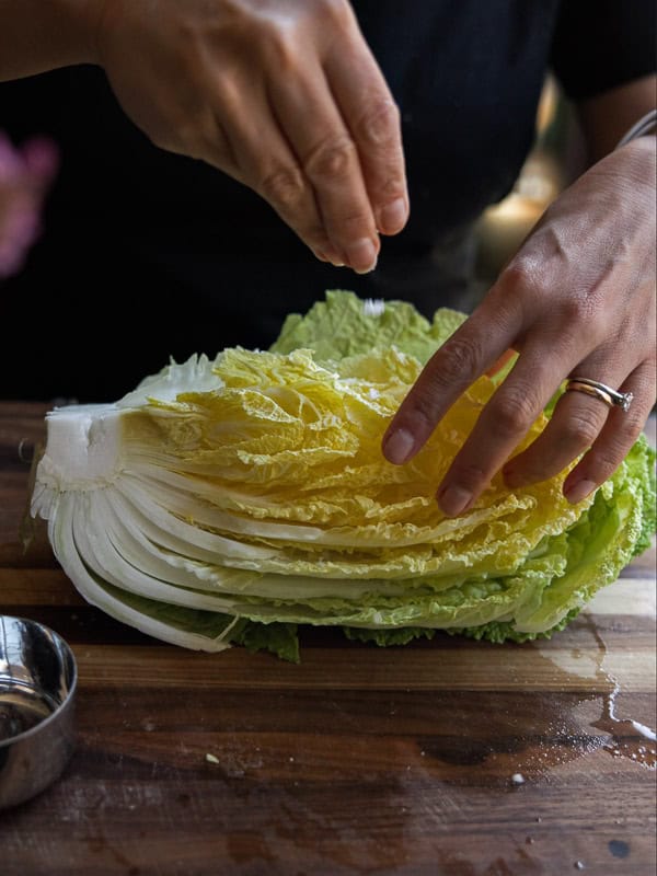 Mom's Easy White Kimchi (Baek-Kimchi) Recipe (백김치). 15 A person sprinkles salt on a halved napa cabbage on a wooden cutting board. A small metal bowl is nearby. The persons hands show a ring and are positioned over the cabbages layered leaves.