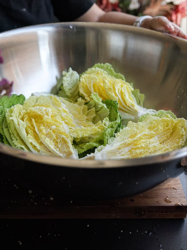 Mom's Easy White Kimchi (Baek-Kimchi) Recipe (백김치). 19 A large stainless steel bowl contains halved pieces of napa cabbage. The bowl is placed on a wooden cutting board, and a person is holding the edge of the bowl. The cabbage leaves are green and pale yellow.