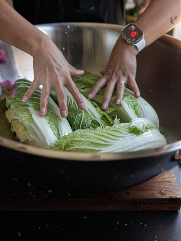 Mom's Easy White Kimchi (Baek-Kimchi) Recipe (백김치). 20 A person wearing a smartwatch is preparing kimchi. They are massaging large cabbage leaves in a metal bowl on a wooden cutting board.