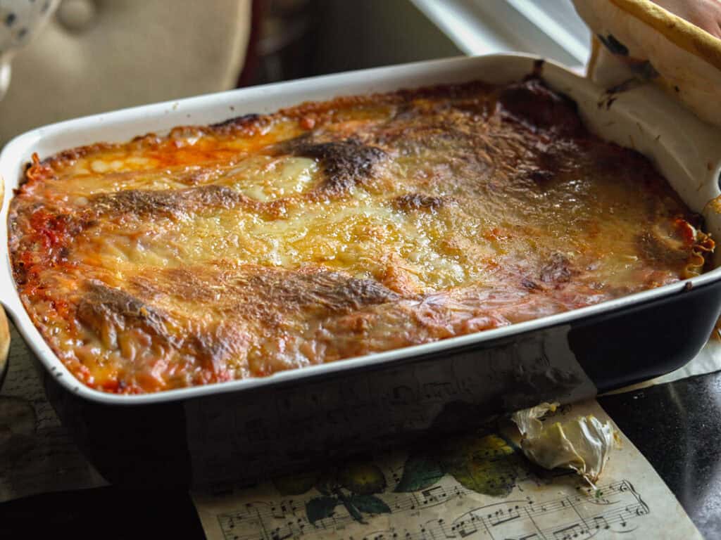 The BEST Vegan Lasagna Recipe - Easy and Delicious. 2 A baked vegan lasagna in a black and white rectangular dish, featuring a golden-brown and slightly crispy cheese topping. It is placed on a table with a printed sheet and a partially visible bread basket nearby.