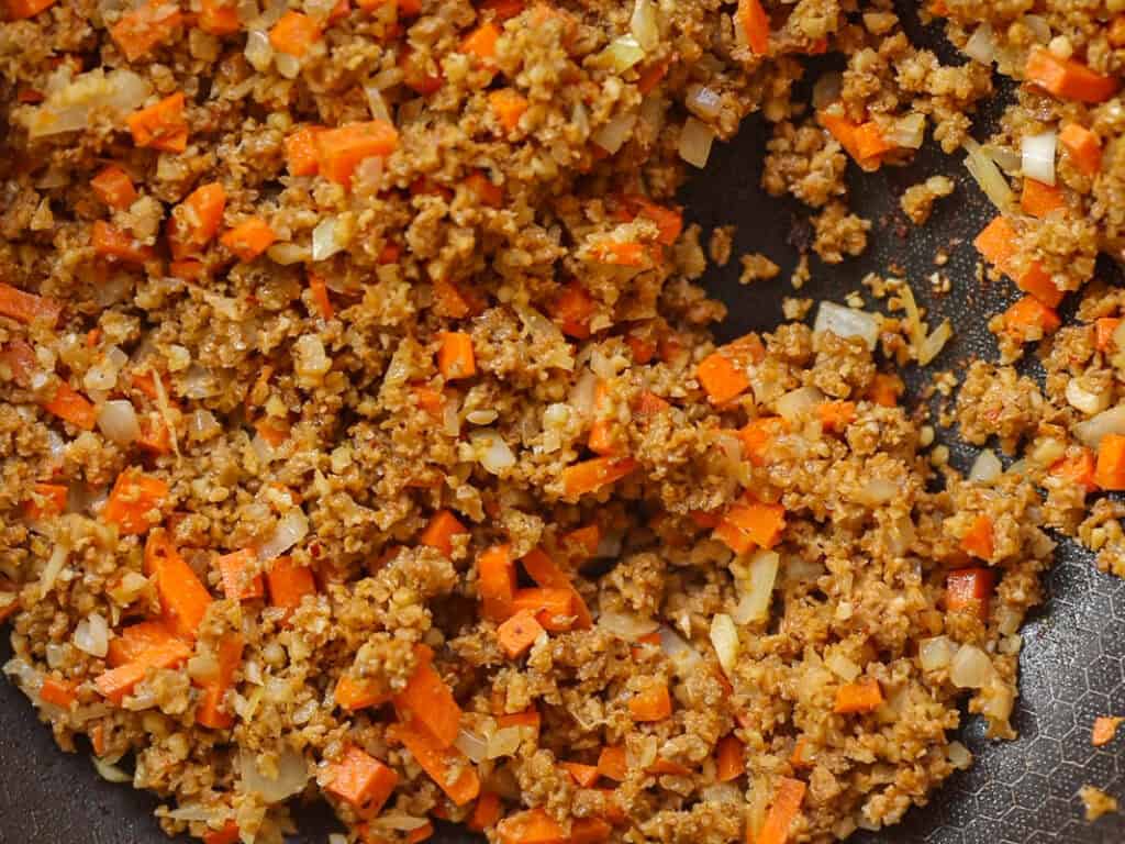 The BEST Vegan Lasagna Recipe - Easy and Delicious. 17 Close-up of sautéed vegetables mixed with cooked ground vegan italian sausage in a non-stick pan. The mixture includes finely chopped carrots and onions, creating a colorful and textured dish.