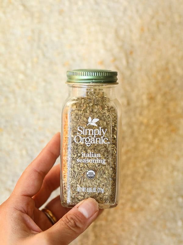 10 Tips for the Perfect Roasted Potatoes Recipe. 13 Hand holding a jar of Simply Organic Italian Seasoning with a metal green lid. Background is a textured, light-colored surface.
