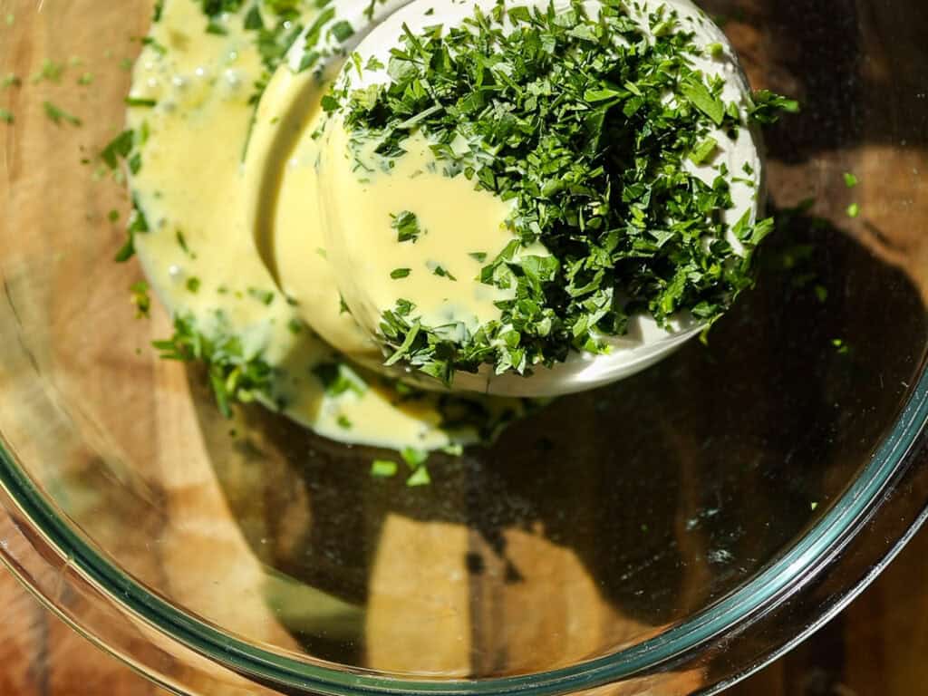 The BEST Vegan Lasagna Recipe - Easy and Delicious. 23 A glass bowl containing a food processor filled with a creamy mixture topped with finely chopped fresh herbs, possibly parsley, placed on a wooden surface. The herbs are vibrant green and the mixture is pale yellow.