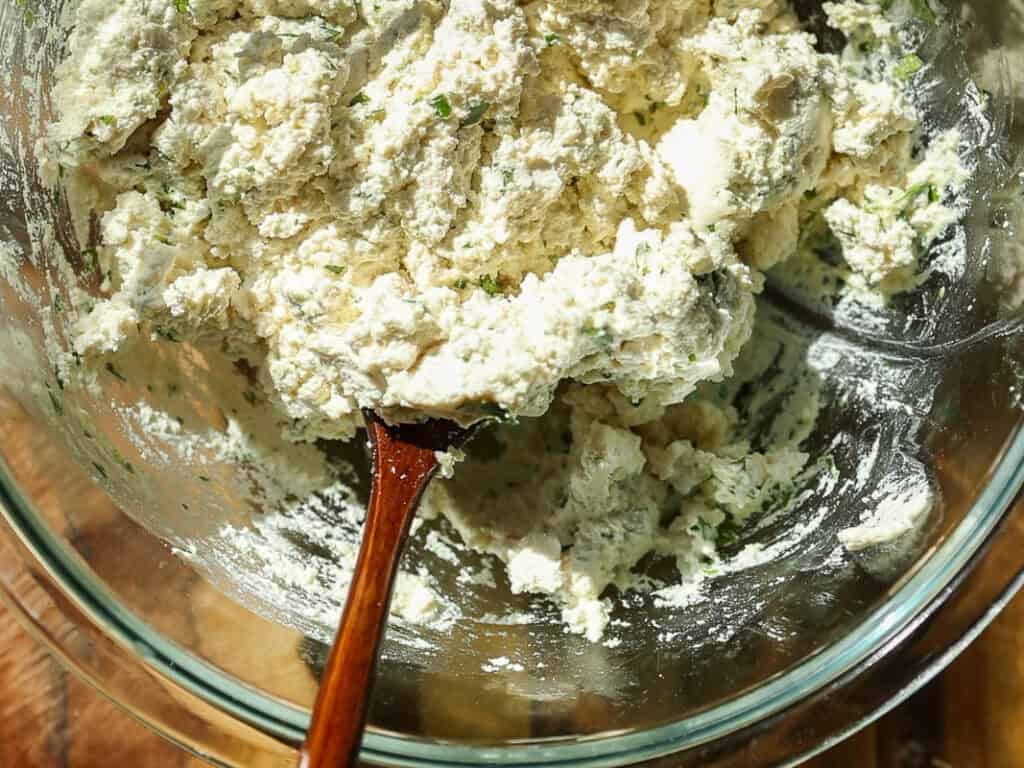 The BEST Vegan Lasagna Recipe - Easy and Delicious. 24 A glass bowl filled with fluffy ricotta cheese mixed with herbs. A wooden spoon is partially submerged in the mixture. The bowl is placed on a wooden surface, and bright sunlight highlights the texture and colors.