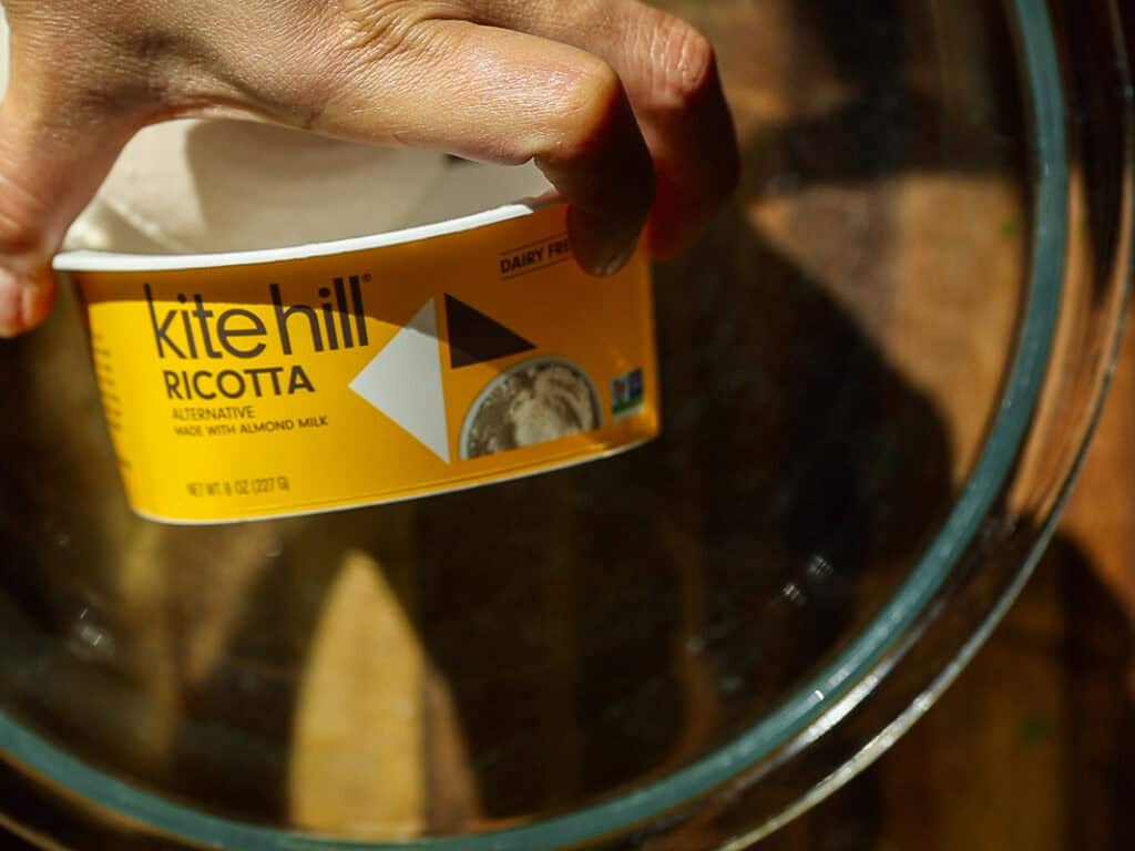 The BEST Vegan Lasagna Recipe - Easy and Delicious. 6 Hand holding a container of Kite Hill dairy-free ricotta made with almond milk, against a wooden background. The container is labeled as an alternative product.
