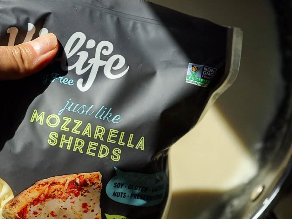 The BEST Vegan Lasagna Recipe - Easy and Delicious. 11 A close-up of a hand holding a black package labeled just like mozzarella shreds from a dairy-free brand. The packaging highlights non-GMO verification and is soy, gluten, lactose, nut, and preservative-free. A pizza image is visible.