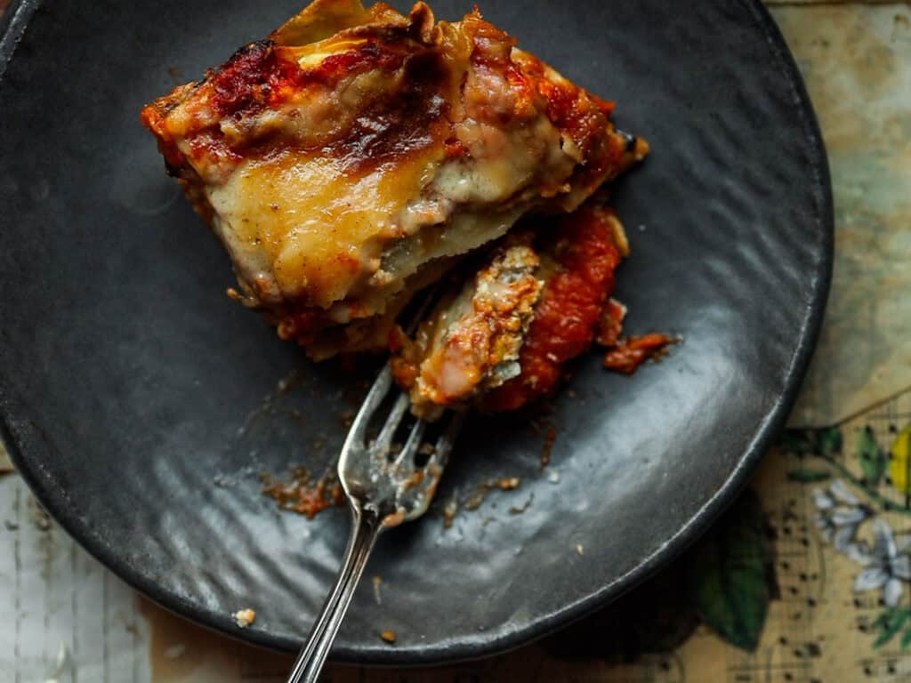 The BEST Vegan Lasagna Recipe - Easy and Delicious. 43 A slice of vegan lasagna is on a dark plate, with a fork partially inserted into it. The lasagna is layered with visible cheese, sauce, and pasta, with a slightly crispy top. It sits on a textured tabletop.