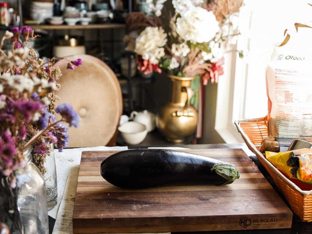The BEST Vegan Lasagna Recipe - Easy and Delicious. 8 An eggplant rests on a wooden cutting board in a rustic kitchen. Behind it, a vase of flowers sits on a table, along with various kitchen items and decor. Sunlight streams through a nearby window, illuminating the scene.