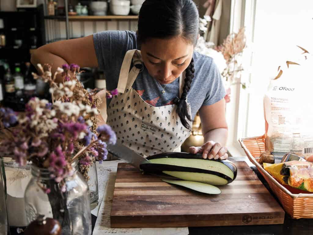 The BEST Vegan Lasagna Recipe - Easy and Delicious. 12 A woman in a blue shirt and apron is preparing food in a kitchen. She is carefully slicing a large eggplant on a wooden cutting board. The kitchen is adorned with dried flowers and various cooking ingredients.