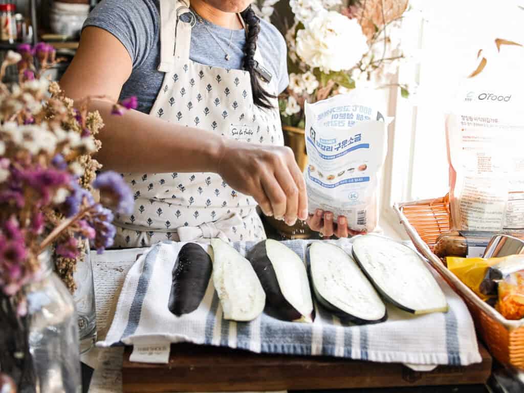 The BEST Vegan Lasagna Recipe - Easy and Delicious. 13 A person in a gray shirt and patterned apron is seasoning sliced eggplants on a towel-covered board. Surrounding the scene are flowers and cooking items, with sunlight streaming in from the side.