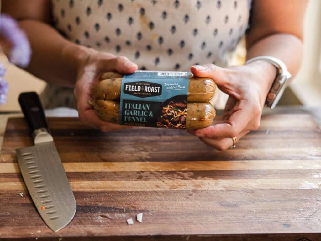 The BEST Vegan Lasagna Recipe - Easy and Delicious. 7 A person holds a package of Field Roast Italian Garlic & Fennel plant-based sausages. A knife is on a wooden cutting board in front of them. The setting appears to be a kitchen.