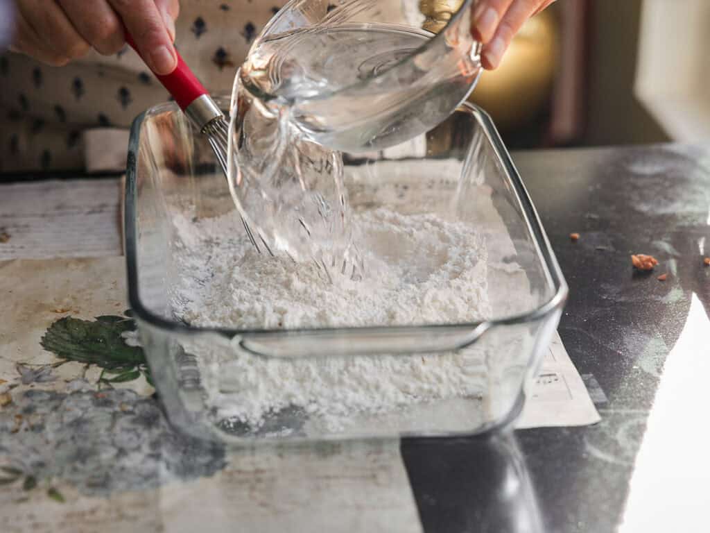 The BEST Vegan Lasagna Recipe - Easy and Delicious. 18 A person pours water from a glass bowl into a rectangular glass dish filled with flour. The scene is set on a table with a floral design.