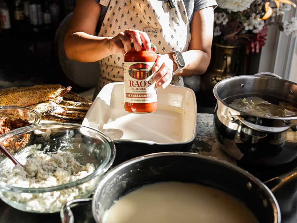 The BEST Vegan Lasagna Recipe - Easy and Delicious. 4 Person preparing a dish by spreading Raos tomato sauce in a rectangular baking dish. Various ingredients like cheese and pasta are arranged around, and theres a pot of white sauce nearby. The setting suggests a cozy kitchen environment.