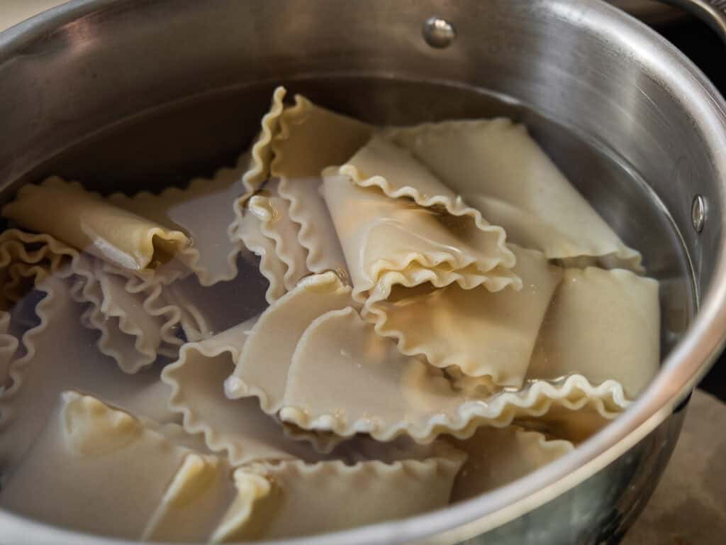 The BEST Vegan Lasagna Recipe - Easy and Delicious. 5 Lasagna noodles are being boiled in a stainless steel pot filled with water. The noodles are folded and partially submerged, with steam rising from the water.