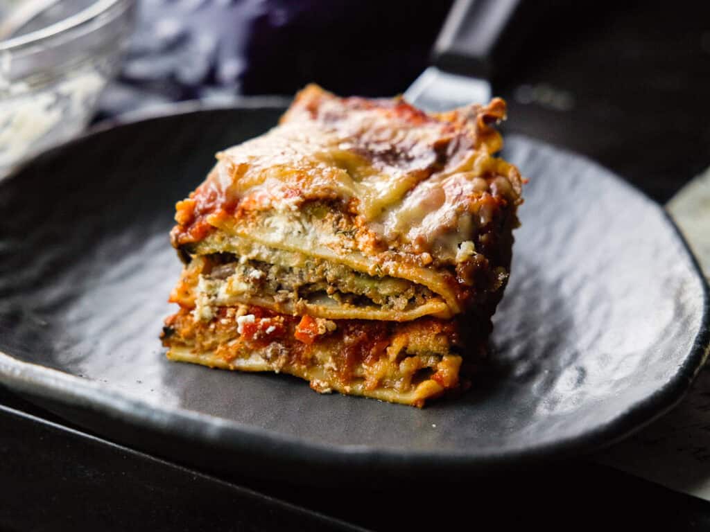The BEST Vegan Lasagna Recipe - Easy and Delicious. 3 A slice of lasagna with layers of pasta, eggplant, cheese, and sauce sits on a dark plate. The lasagna is golden and slightly crispy on top, with a rich, savory filling visible between the layers.