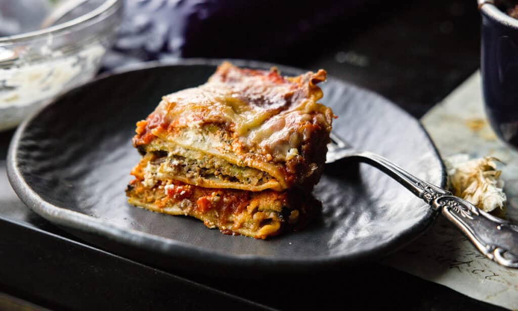 The BEST Vegan Lasagna Recipe - Easy and Delicious. 1 A close-up of a piece of vegan lasagna on a dark plate, with layers of pasta, rich tomato sauce, and melted cheese. A fork rests beside it. The setting has a rustic and cozy feel.