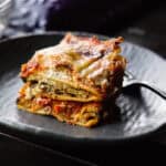 30+ AMAZING VEGAN Holiday Recipes Everyone's gonna Love. 11 A close-up of a slice of vegan lasagna on a dark, round plate. The lasagna has visible layers of pasta, eggplant, tomato sauce, and melted cheese. A fork is placed on the plate next to the lasagna.