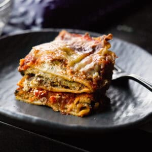The BEST Vegan Lasagna Recipe - Easy and Delicious. 47 A close-up of a slice of vegan lasagna on a dark, round plate. The lasagna has visible layers of pasta, eggplant, tomato sauce, and melted cheese. A fork is placed on the plate next to the lasagna.