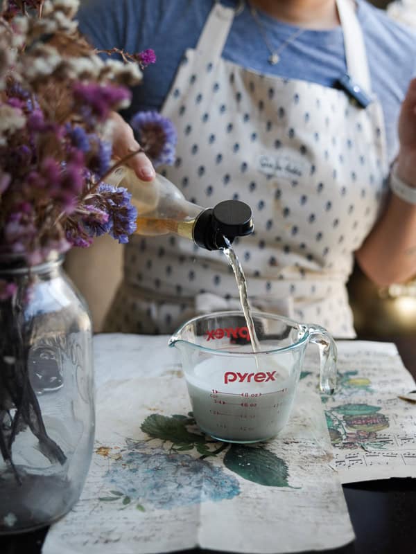 Perfect Roasted Garlic Vegan Dinner Rolls Recipe. 9 A person wearing a patterned apron pours liquid from a bottle into a Pyrex measuring cup filled with oat milk. Flowers in a glass jar are visible on the left. The scene is set on a table with floral paper.