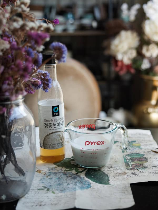 Perfect Roasted Garlic Vegan Dinner Rolls Recipe. 6 A glass bottle of rice vinegar stands beside a Pyrex measuring cup containing oat milk. They are on a table covered with floral-patterned paper, next to a vase of dried flowers. Blurred floral decor is visible in the background.