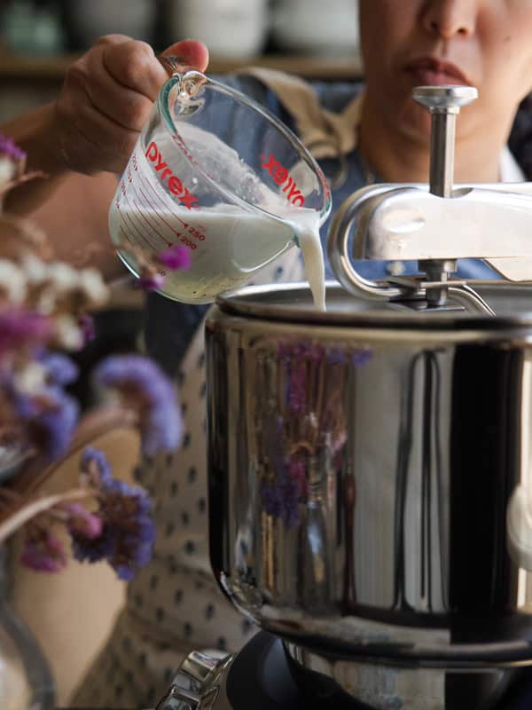 Perfect Roasted Garlic Vegan Dinner Rolls Recipe. 15 A person pouring vegan buttermilk from a measuring cup into a shiny metal stand mixer. Purple flowers are blurred in the foreground, and the person is wearing a white apron with a blue shirt.
