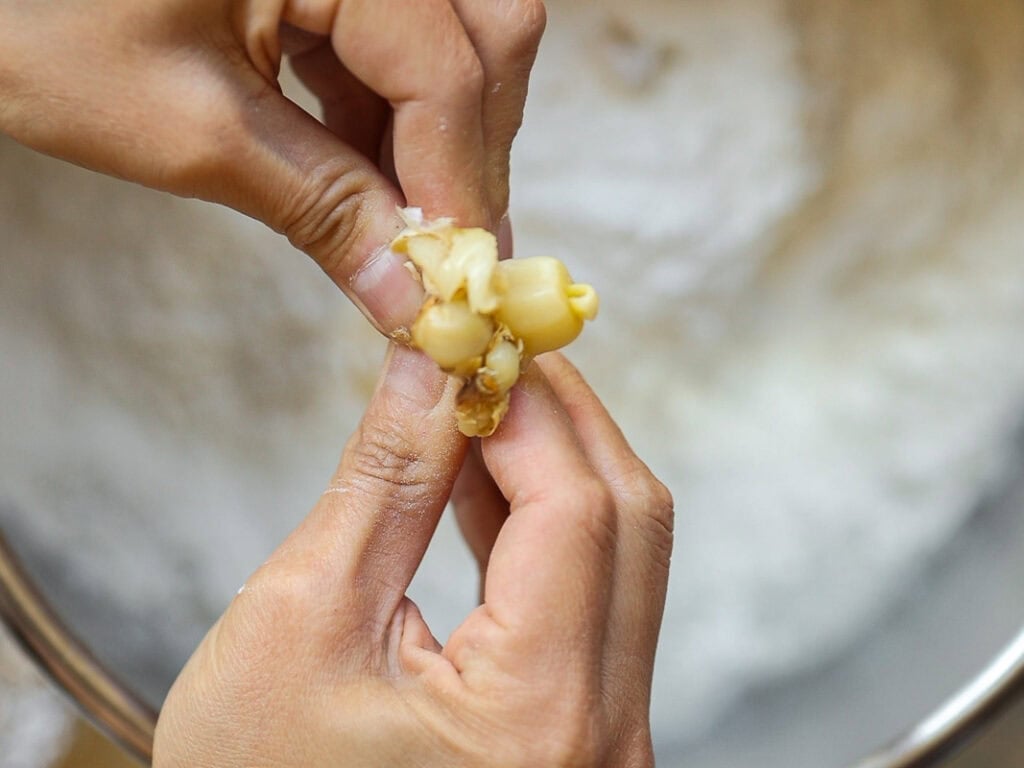 Perfect Roasted Garlic Vegan Dinner Rolls Recipe. 12 Close-up of hands squeezing out pieces of roasted garlic in front of a bowl with a white powdered substance, possibly flour.