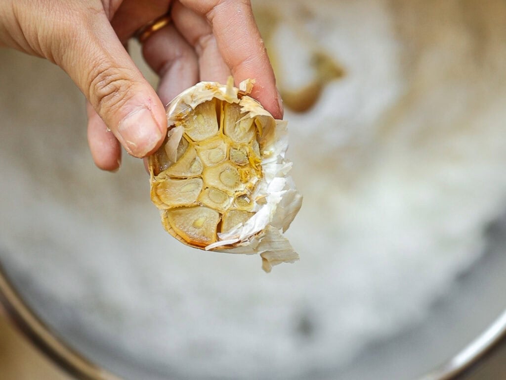 Perfect Roasted Garlic Vegan Dinner Rolls Recipe. 4 A hand holds a roasted garlic bulb with golden-brown cloves visible. The background is blurred, highlighting the garlic in the foreground.