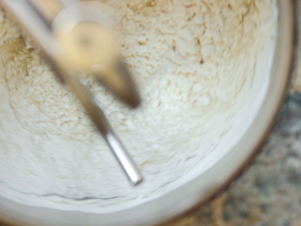 Perfect Roasted Garlic Vegan Dinner Rolls Recipe. 14 A close-up of a mixer in a mixing bowl with a dough being stirred. The motion blur indicates the mixer is in use, blending the ingredients. The bowl is partially visible against a speckled countertop.