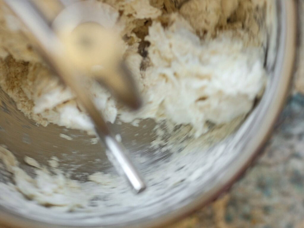Perfect Roasted Garlic Vegan Dinner Rolls Recipe. 16 Close-up of dough in a metal mixing bowl, partially mixed by a silver dough hook. The dough is light in color and appears to be in the process of being kneaded. The background shows a speckled countertop.
