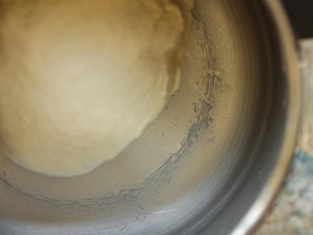 Perfect Roasted Garlic Vegan Dinner Rolls Recipe. 19 A close-up of dough in a mixing bowl. The dough is smooth and pale in color, partially stuck to the side of the stainless steel bowl, suggesting its being prepared for baking.