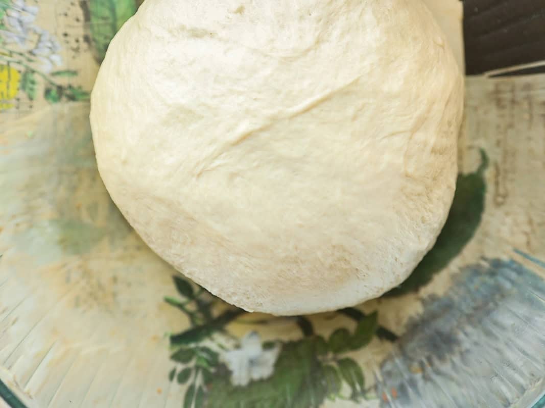 Perfect Roasted Garlic Vegan Dinner Rolls Recipe. 21 A ball of raw dough sits on a patterned glass plate. The dough is smooth and pale, resting atop a colorful floral design printed on the plate, which provides a contrasting backdrop.