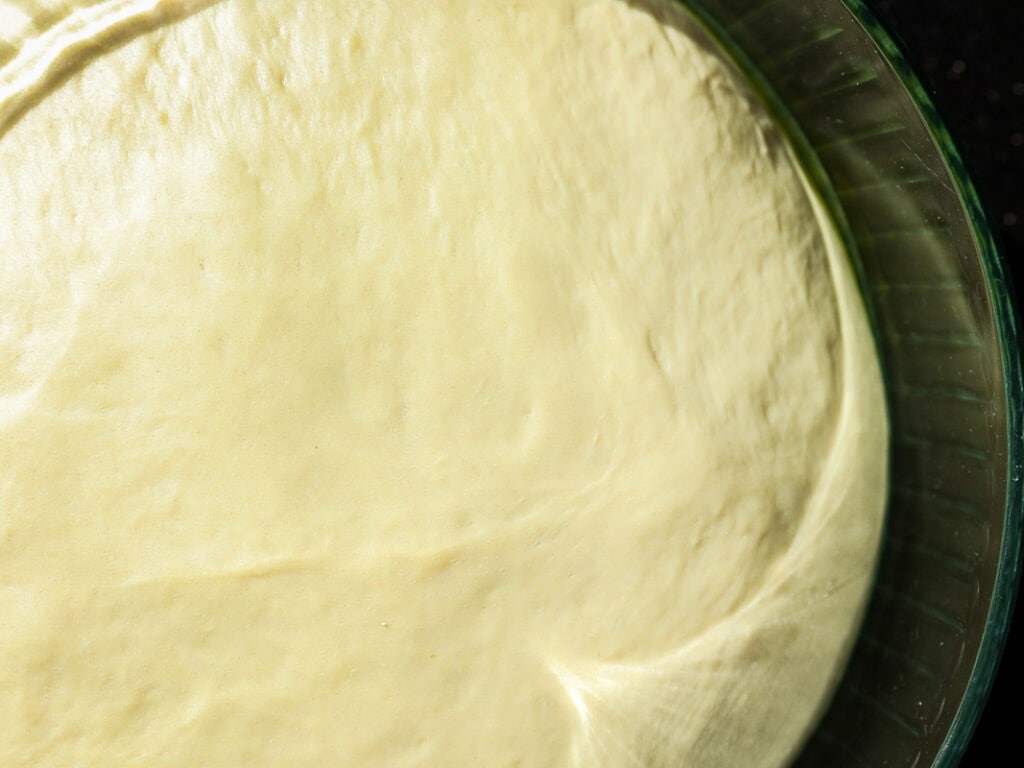 Perfect Roasted Garlic Vegan Dinner Rolls Recipe. 22 A close-up of smooth, risen dough in a glass bowl, ready for baking. The surface is slightly glossy, with an even texture, illuminated by soft light.