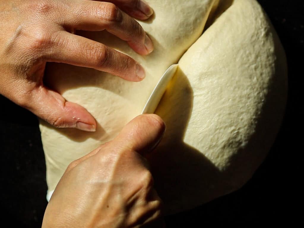 Perfect Roasted Garlic Vegan Dinner Rolls Recipe. 24 Hands seen from above knead and slice a large dough ball with a knife on a dark surface, highlighting the texture and action in natural light.