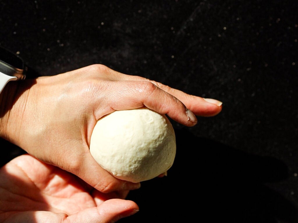 Perfect Roasted Garlic Vegan Dinner Rolls Recipe. 26 A person shaping dough into a round ball with their hands against a dark background, indicating a baking or cooking process.