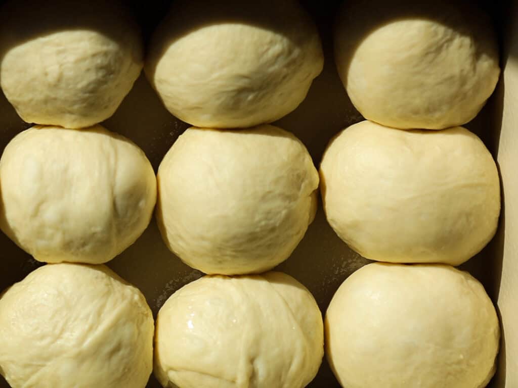 Perfect Roasted Garlic Vegan Dinner Rolls Recipe. 30 Eight round balls of dough arranged in rows, resting on a surface. The doughs surface is smooth and slightly shiny, illuminated by natural light.
