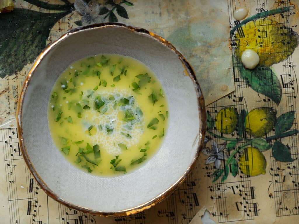 Perfect Roasted Garlic Vegan Dinner Rolls Recipe. 32 A bowl of vegan egg wash mixture with chopped herbs on a vintage sheet music page featuring lemon illustrations. The edges of the bowl are gold-rimmed, and the surface below includes splashes of the mixture.