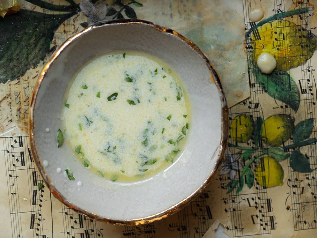 Perfect Roasted Garlic Vegan Dinner Rolls Recipe. 33 A bowl of vegan "egg wash" with chopped herbs, placed on a vintage music sheet with lemon illustrations. A few splatters of soup are visible around the bowl.